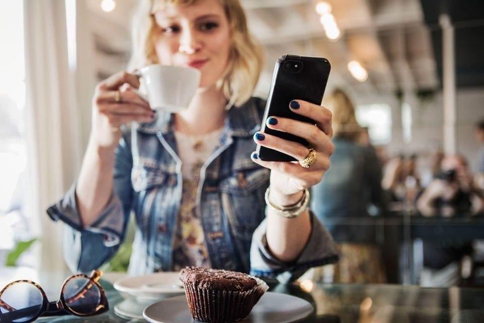 Woman seated at a restaurant sips from a mug while looking at her phone.
