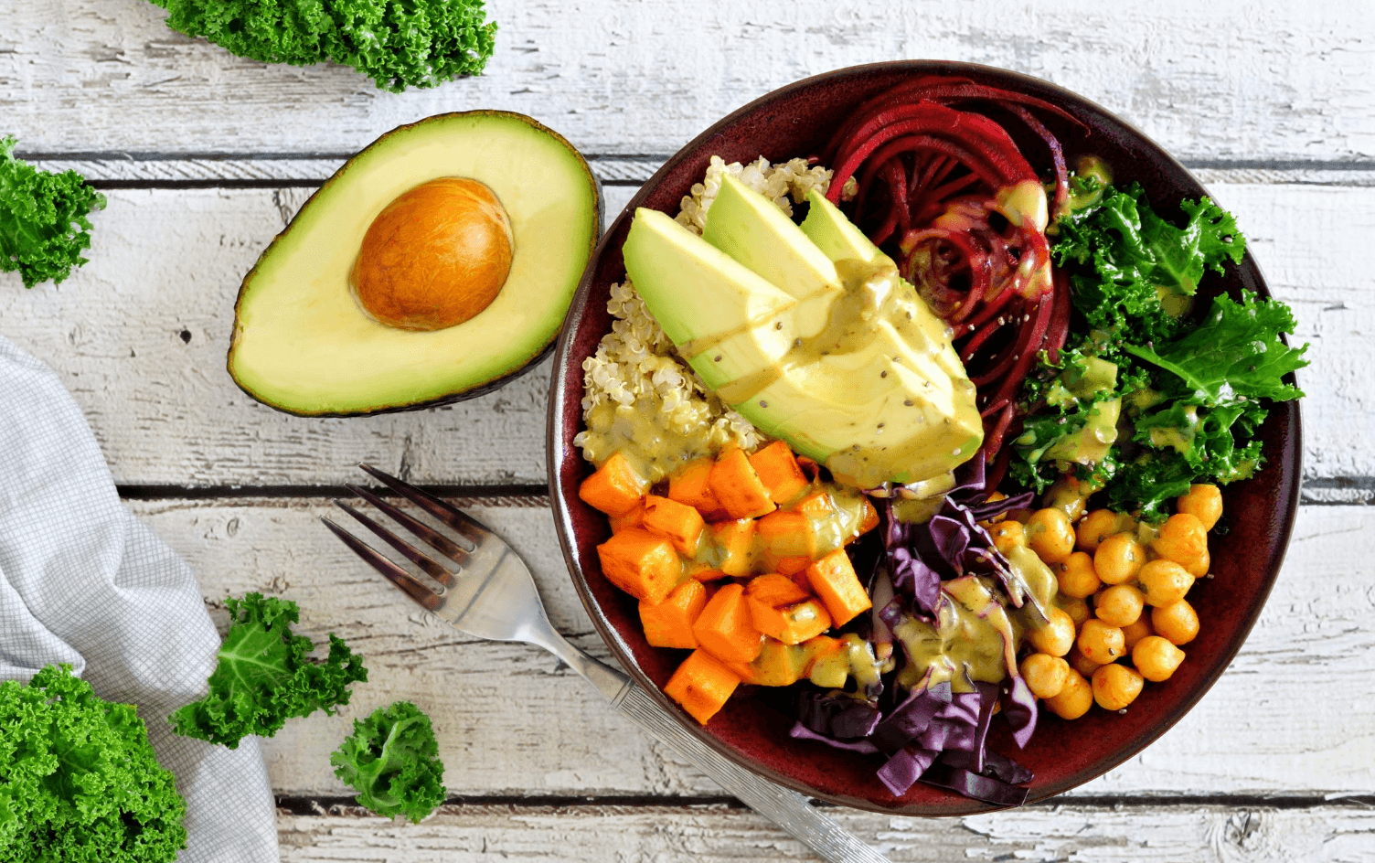 Overhead view of a bowl filled with sweet potato, quinoa, cabbage, chickpeas, beets, and greens, with half an avocado and a fork lying to the side.